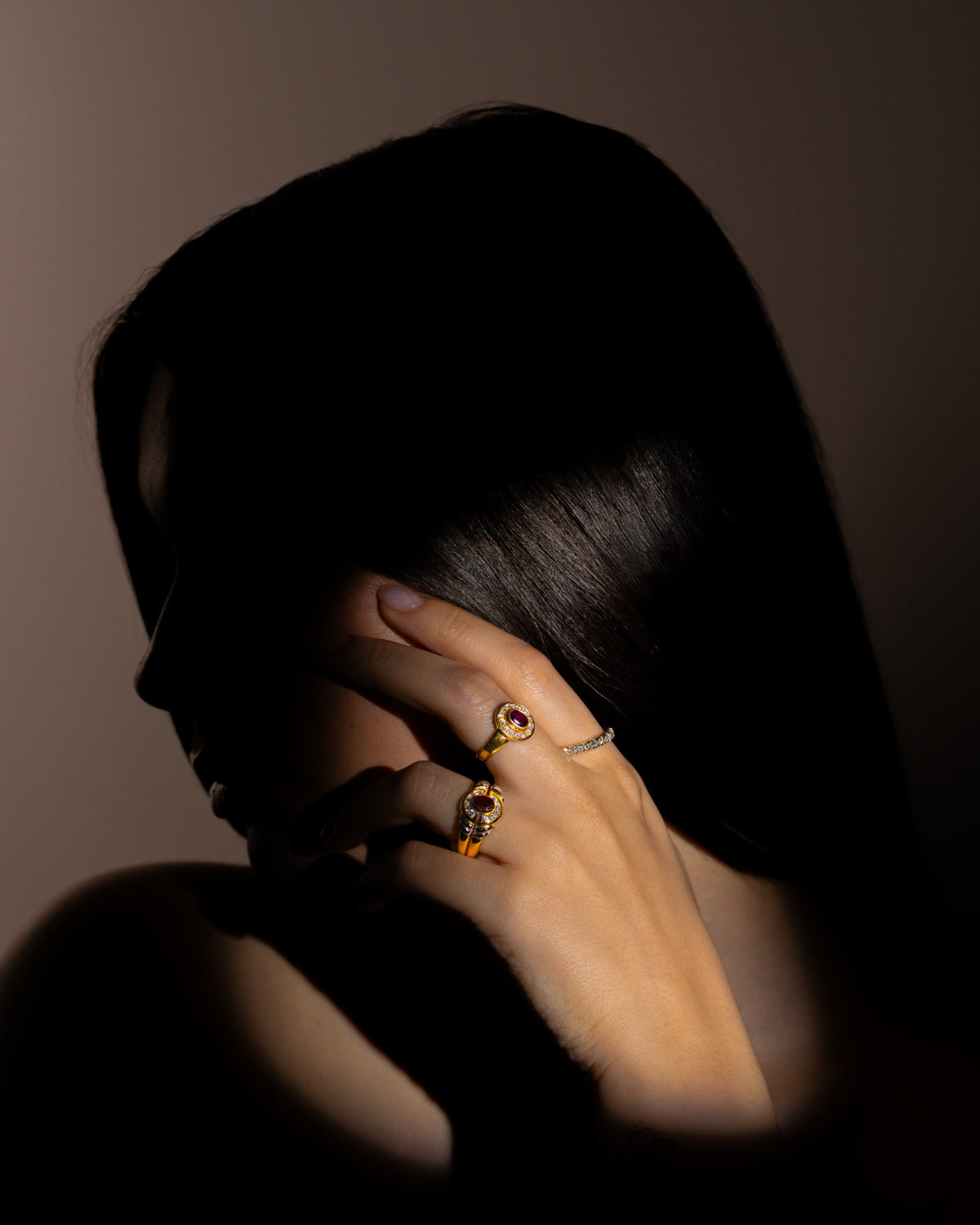 Anello con rubino ovale e corona di diamanti in oro giallo indossato al dito, mano tra i capelli, con altri anelli