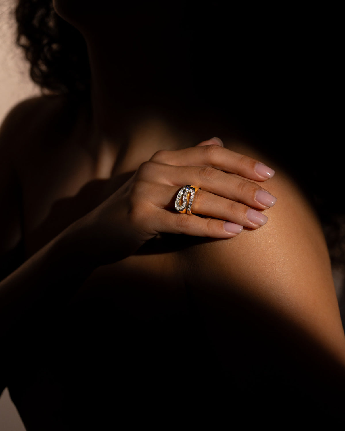 Anello S di diamanti in oro 18 carati indossato al dito, mano appoggiata sulla spalla in luce calda