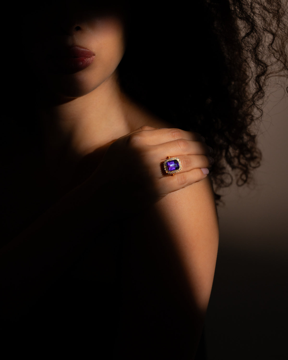 Anello ametista e diamanti in oro giallo indossato al dito, mano appoggiata sulla spalla, luce di taglio su fondo scuro
