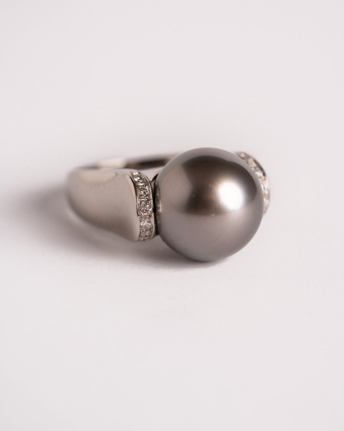Anello con perla di Tahiti in oro bianco visto di lato, piccole pietre bianche sulle spalle, still life su fondo chiaro