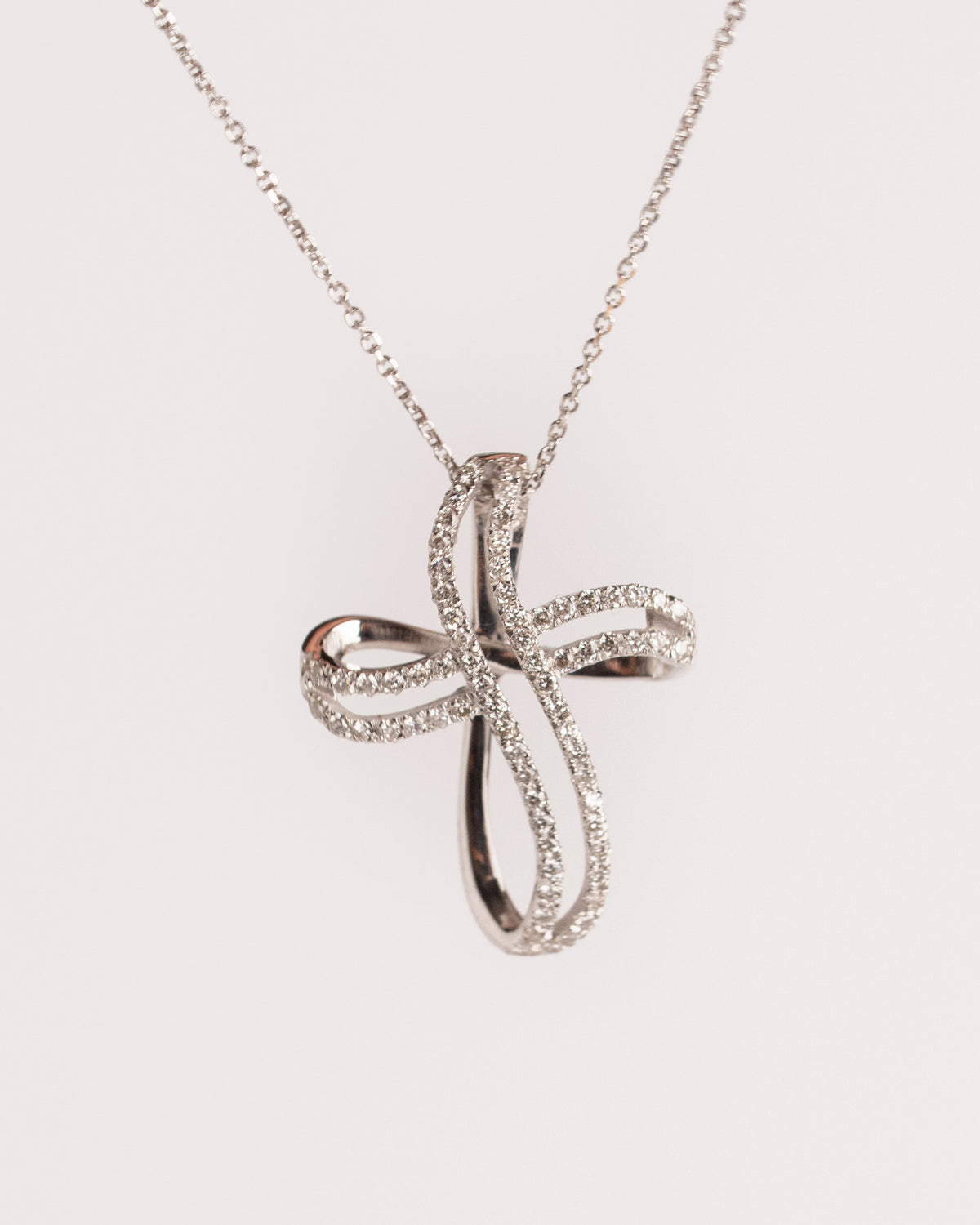 Croce contemporanea in oro bianco con pavé di diamanti e bracci curvi intrecciati, still life su fondo chiaro